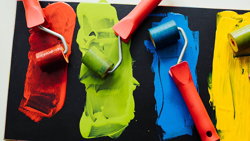 Vier Farbroller mit roten Griffen auf schwarzem Untergrund, jeweils mit roter, grüner, blauer und gelber Farbe.