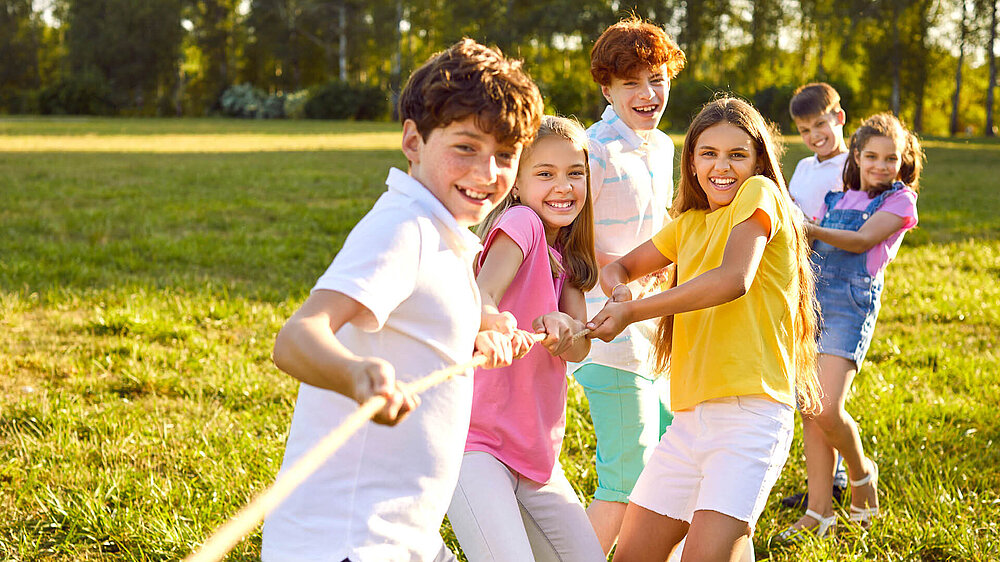Sechs lachende Kinder spielen Tauziehen auf einer Wiese im Sonnenschein.