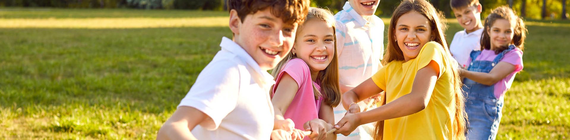 Sechs lachende Kinder spielen Tauziehen auf einer Wiese im Sonnenschein.