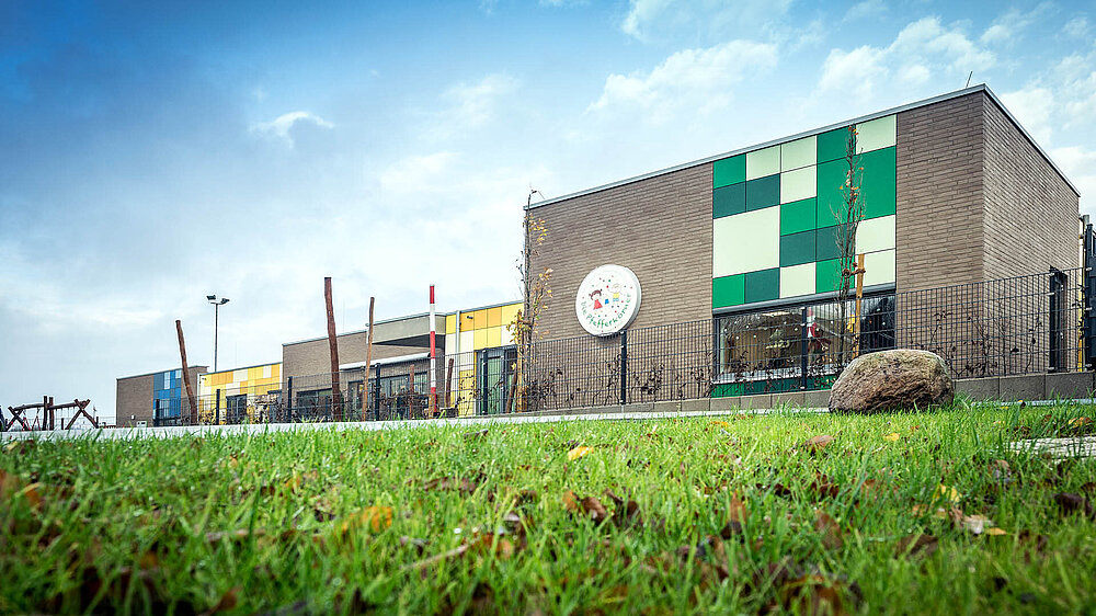 Modernes Gebäude der Kindertagesstätte Die Pfefferkörner mit farbigen Fassadenelementen und Spielplatz im Hintergrund.