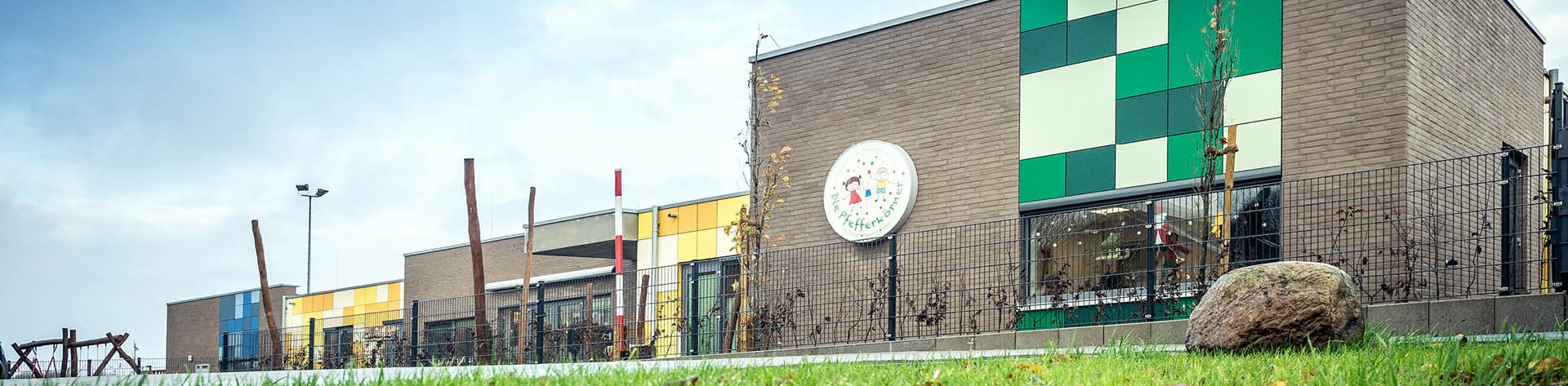 Modernes Gebäude der Kindertagesstätte Die Pfefferkörner mit farbigen Fassadenelementen und Spielplatz im Hintergrund.