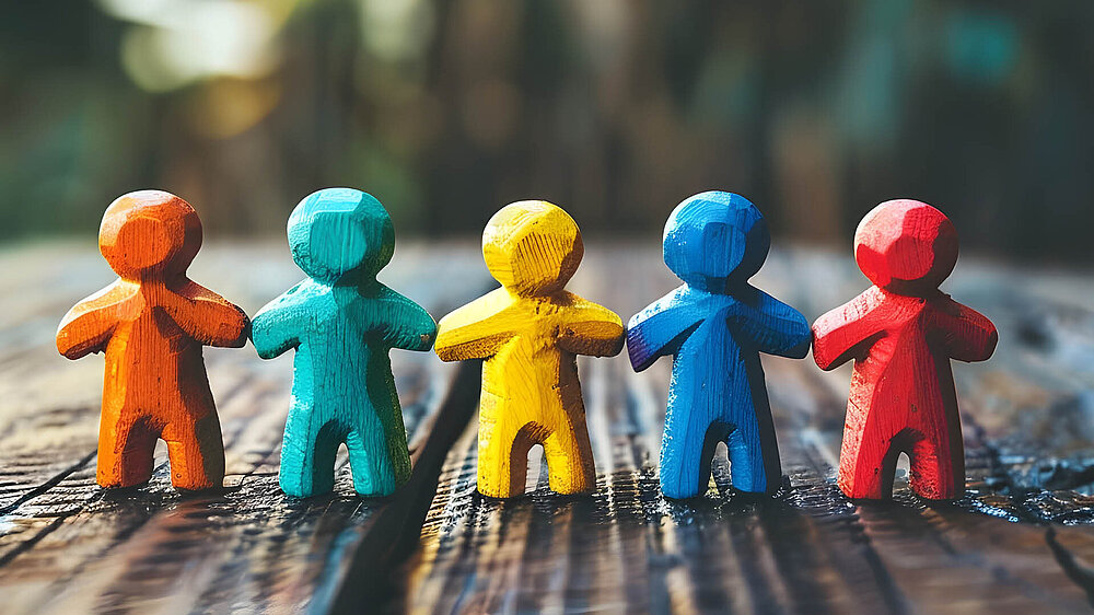 Fünf bunte Holzfiguren in Orange, Türkis, Gelb, Blau und Rot stehen nebeneinander auf einer Holzoberfläche.