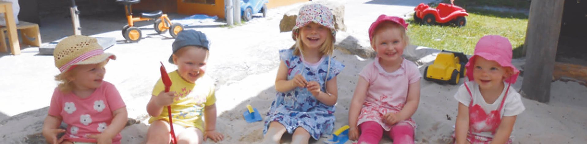 Fünf Kinder sitzen bei sonnigen Wetter draußen in einem Sandkasten vor dem Gebäude der Kindertagesstätte Regenbogen.