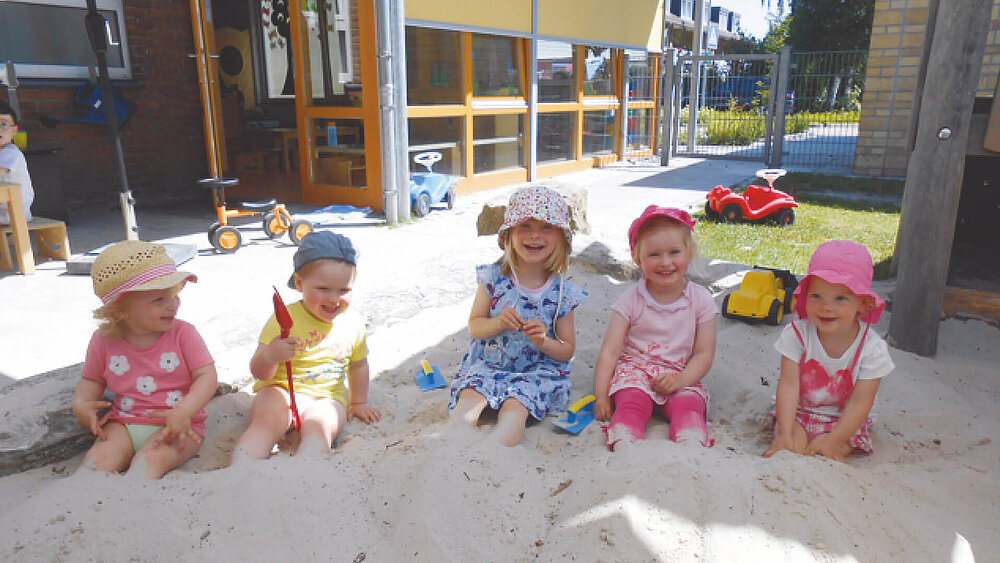 Fünf Kinder sitzen bei sonnigen Wetter draußen in einem Sandkasten vor dem Gebäude der Kindertagesstätte Regenbogen.