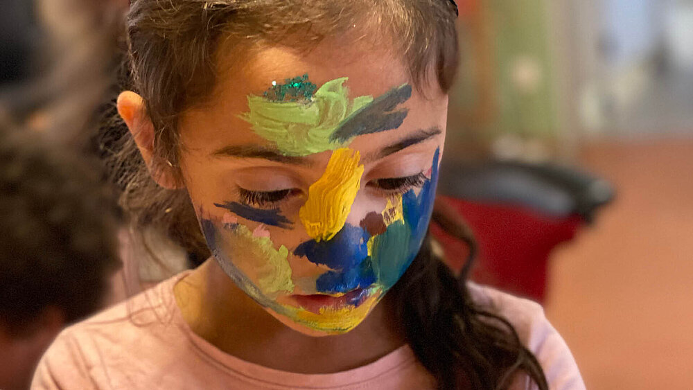 Ein Mädchen mit bemaltem Gesicht hält einen Pinsel in der Hand und malt sich selbst die Hände an.