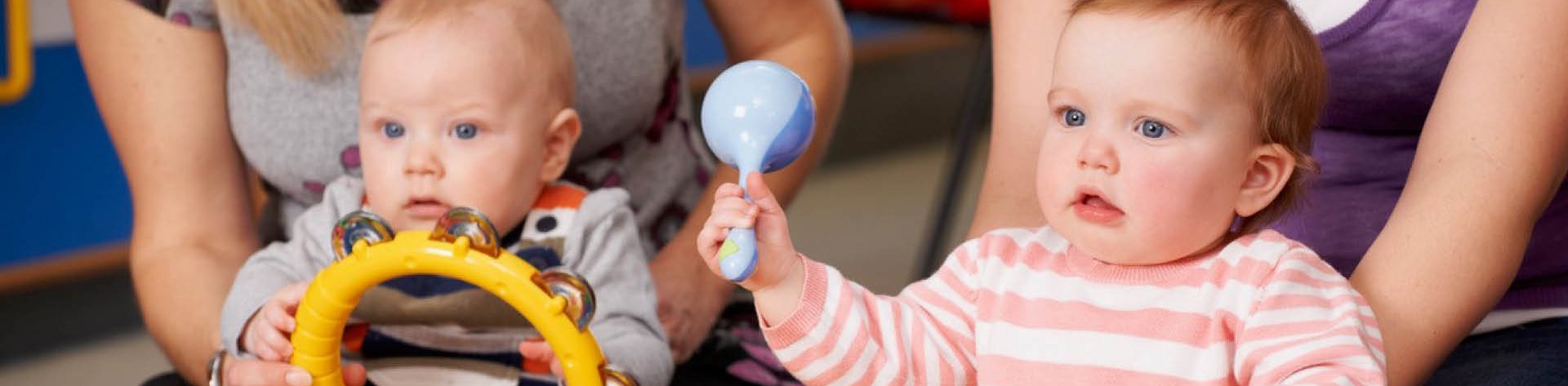 Zwei Babys sitzen auf dem Boden, jedes mit einem Erwachsenem im Hintergrund. Die Babys halten Spielzeuge in den Händen. 