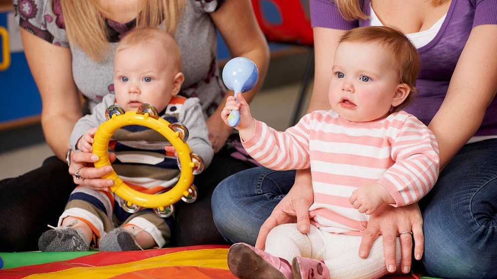 Zwei Babys sitzen auf dem Boden, jedes mit einem Erwachsenem im Hintergrund. Die Babys halten Spielzeuge in den Händen. 
