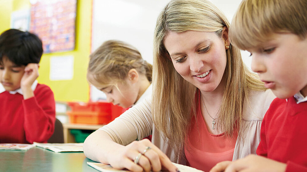 Pädagogische Fachkraft unterstützt einen Schüler im Klassenzimmer beim Lernen, im Hintergrund sind zwei weitere Schüler zu erkennen.
