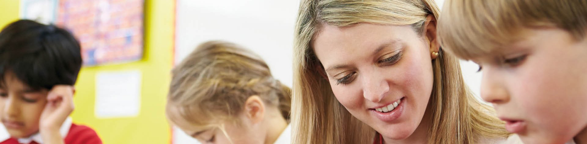Pädagogische Fachkraft unterstützt einen Schüler im Klassenzimmer beim Lernen, im Hintergrund sind zwei weitere Schüler zu erkennen.