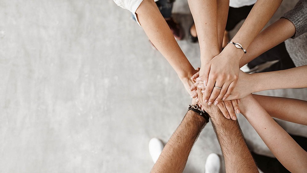 Mehrere Hände verschiedener Personen übereinandergelegt als Zeichen von Teamarbeit und Zusammenhalt. 