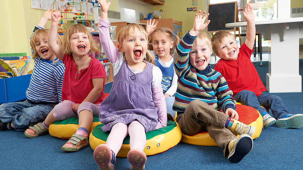 Sechs Kinder sitzen auf dem Boden in einem Raum einer Kindertagesstätte und melden sich mit glücklichen Gesichtern.