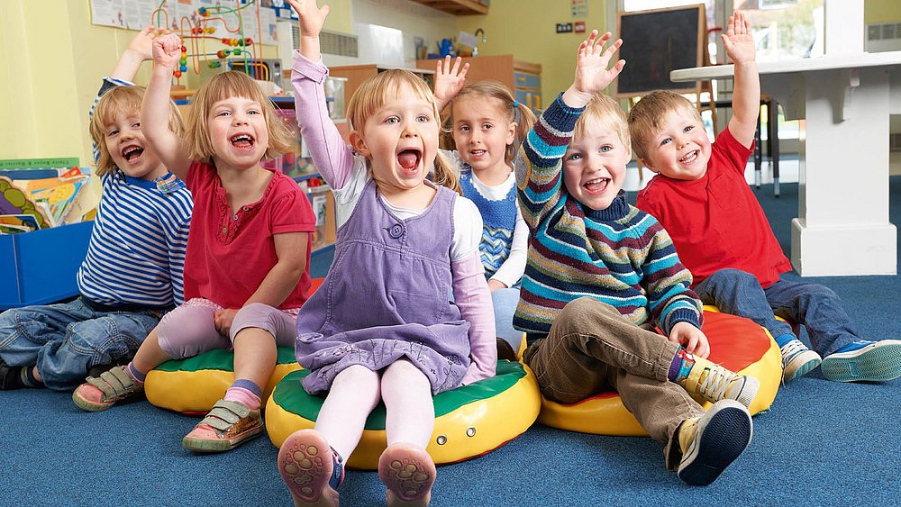Sechs Kinder sitzen auf dem Boden in einem Raum einer Kindertagesstätte und melden sich mit glücklichen Gesichtern.