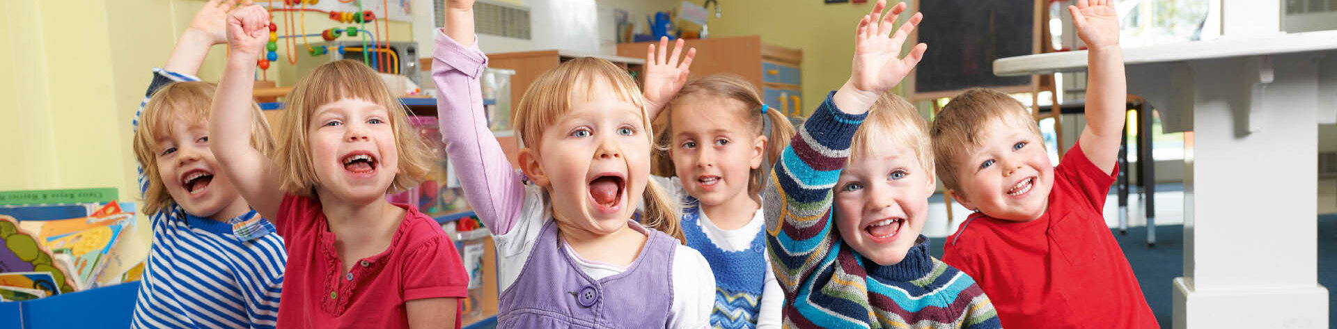 Sechs Kinder sitzen auf dem Boden in einem Raum einer Kindertagesstätte und melden sich mit glücklichen Gesichtern.