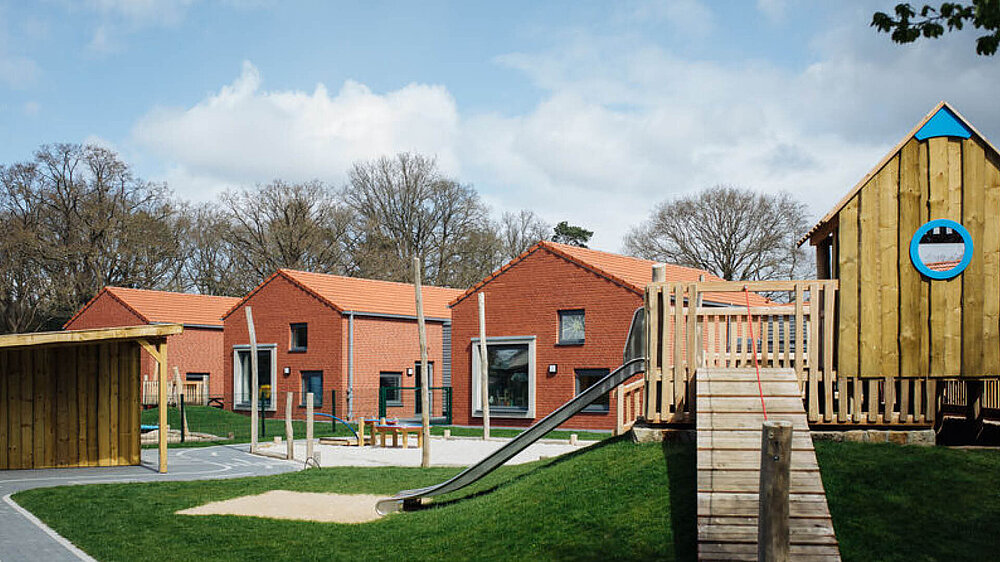 Gebäude der Kindertagesstätte Die Spürnasen mit roten Backsteinhäusern, im Vordergrund ist ein Spielplatz mit Spielhaus und Rutsche zu sehen.