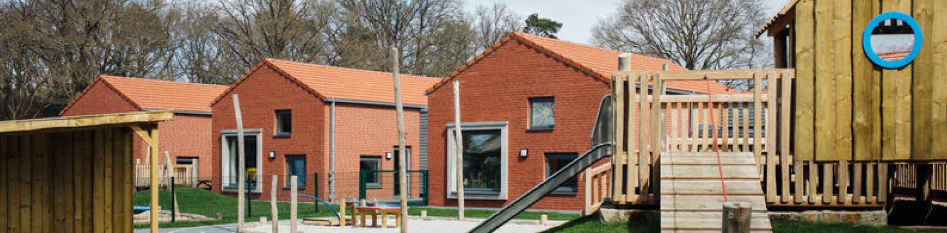 Gebäude der Kindertagesstätte Die Spürnasen mit roten Backsteinhäusern, im Vordergrund ist ein Spielplatz mit Spielhaus und Rutsche zu sehen.
