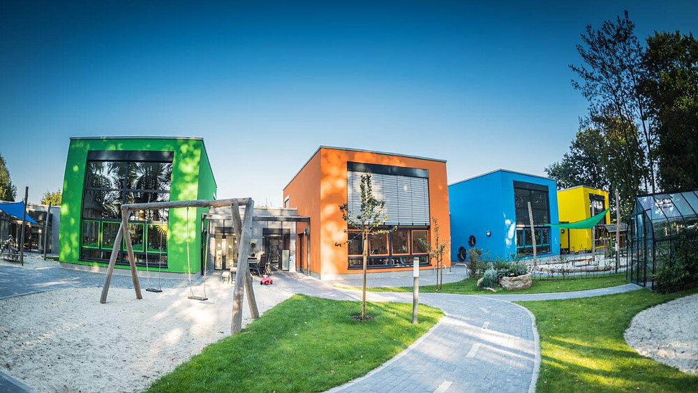 Buntes Gebäude der Kindertagesstätte Kinder(T)räume in den Farben Grün, Orange, Blau und Gelb mit einem Sandplatz und einer Schaukel im Vordergrund.