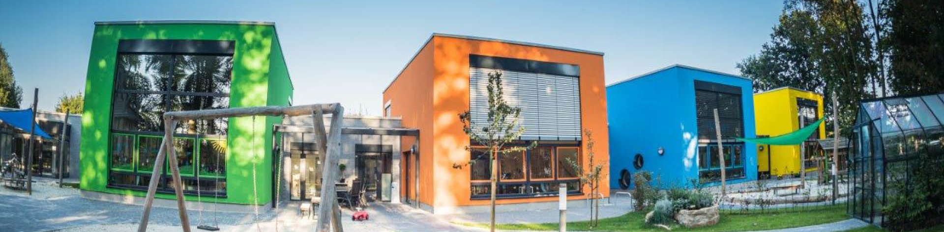 Buntes Gebäude der Kindertagesstätte Kinder(T)räume in den Farben Grün, Orange, Blau und Gelb mit einem Sandplatz und einer Schaukel im Vordergrund.